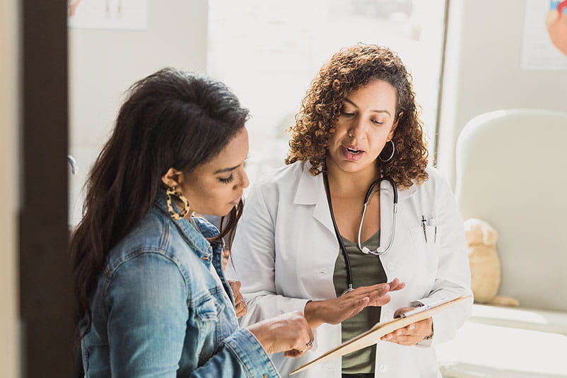 A female patient speaks with a female doctor in a white lab coat.