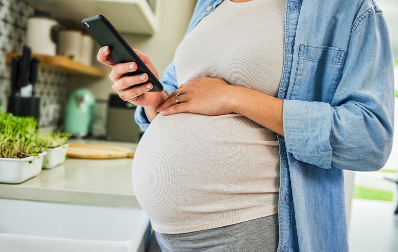 A pregnant woman has a hand on her belly and holds a smartphone.