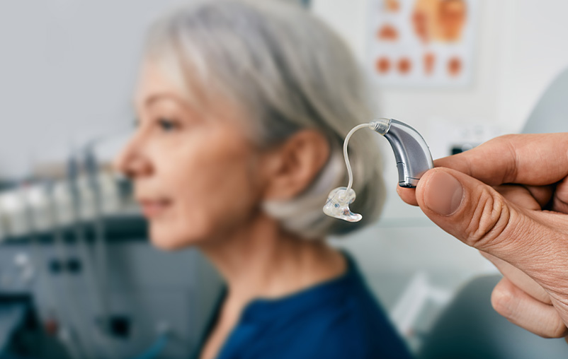 A hand holding a hearing aid. An older woman sits in the background.