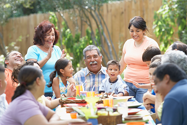 Family is outside sitting at the dinner table. Every is laughing and facing the grandfather.