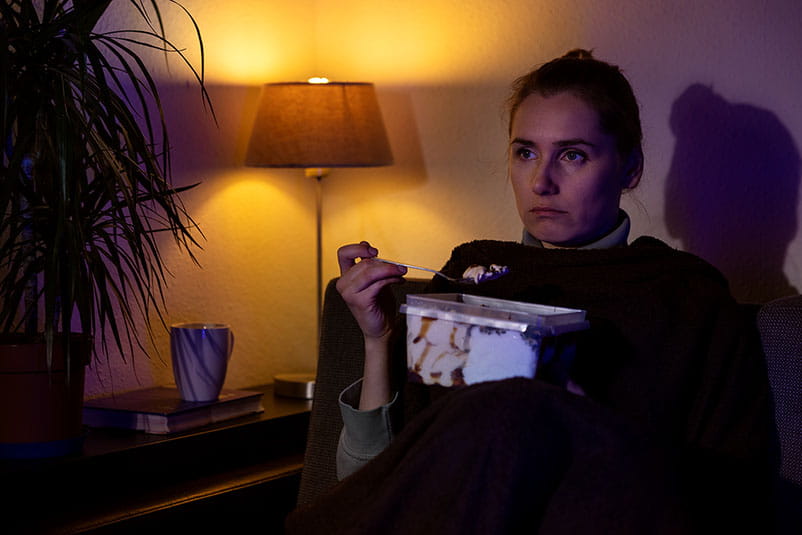 woman eating ice cream from container while sitting on couch and watching tv at home