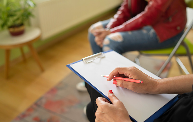 A close-up of a woman holding a pen and clipboard, seated next to a young woman.