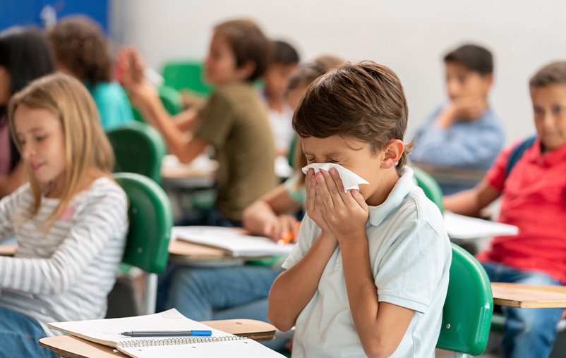 A boy blows his nose sitting at a desk in a classroom