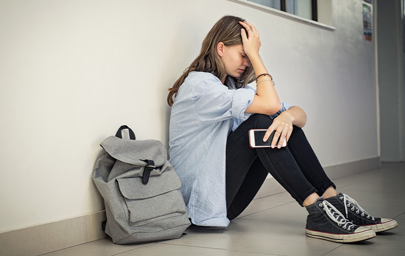 A teenage girl sits on the floor holding her phone with her head in her hand; she looks upset.