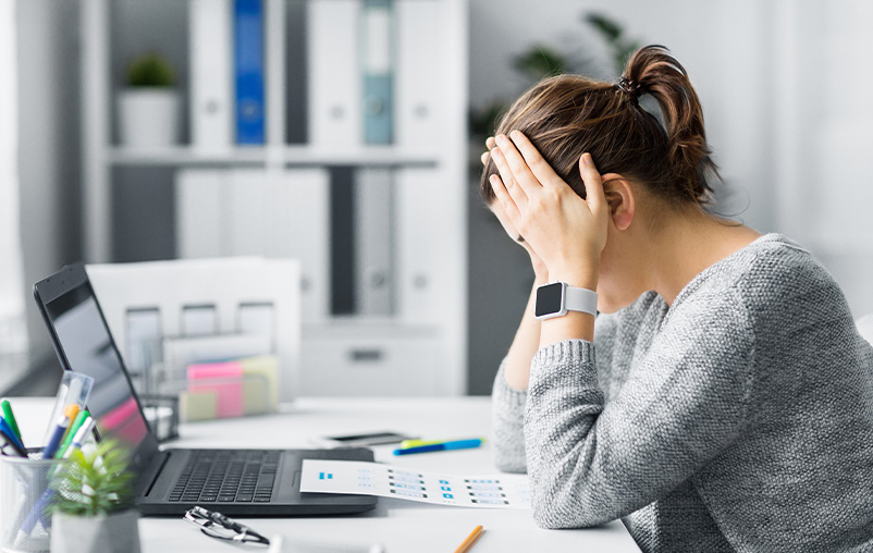 A woman sits at a desk with her head in her hands, looking stressed.