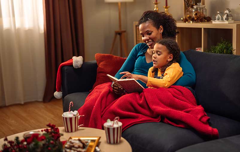 Mother reading a book to her daughter while they are sitting covered in blanket on the sofa during Christmas holidays