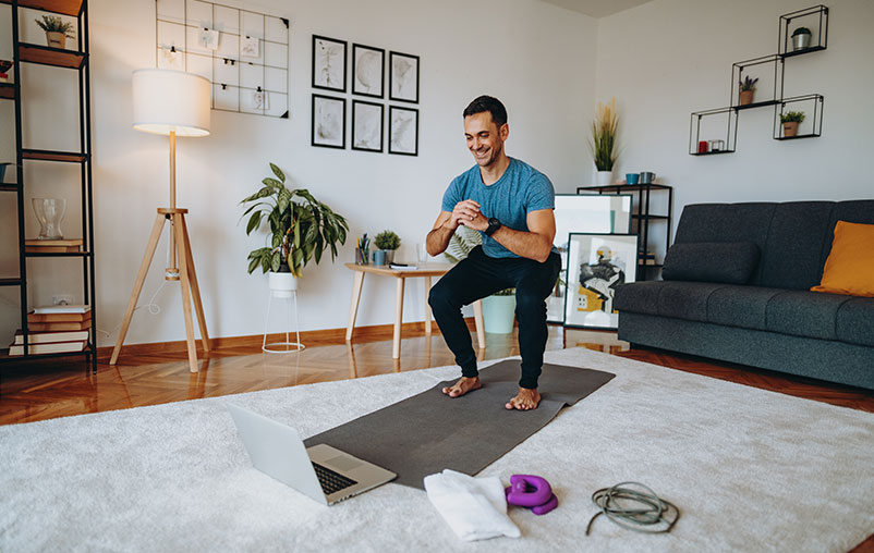 Man in his living room doing squats with exercise equipment near him while viewing a laptop.