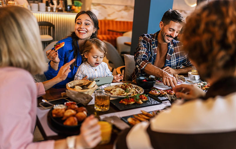 A family sharing a meal at a cozy restaurant, laughing and enjoying food while a child watches a tablet at the table.